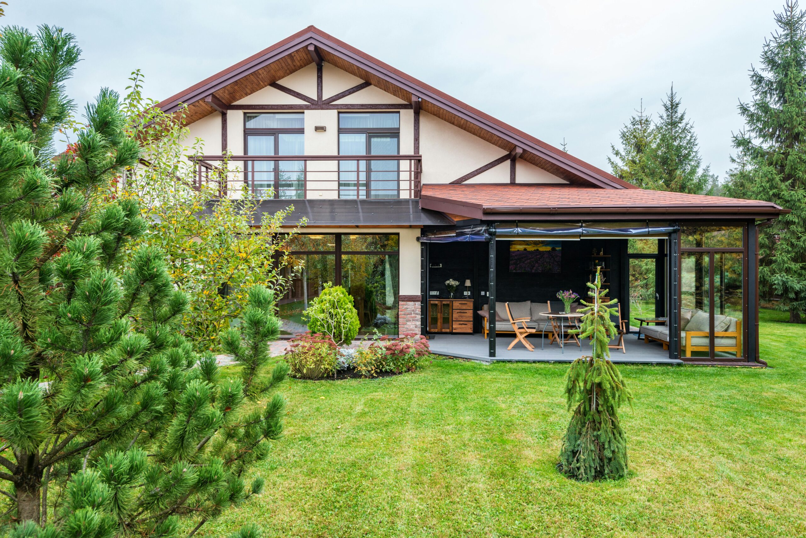 Exterior of contemporary residential house with balcony and cozy terrace with couch near trees on green yard on daytime